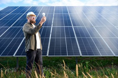 Üniformalı mühendis. Güneş istasyonu yakınlarında koruyucu kasklı. Elinde ampul var. Denetmen güneş çiftliğindeki güneş panellerini kontrol ediyor. Güneş enerjisi ve yenilenebilir enerji. Ön manzara. Yeşil enerji kavramı.