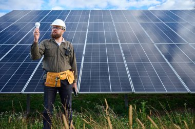 Üniformalı mühendis. Güneş istasyonu yakınlarında koruyucu kasklı. Elinde ampul var. Denetmen güneş çiftliğindeki güneş panellerini kontrol ediyor. Güneş enerjisi ve yenilenebilir enerji. Ön manzara. Yeşil enerji kavramı.