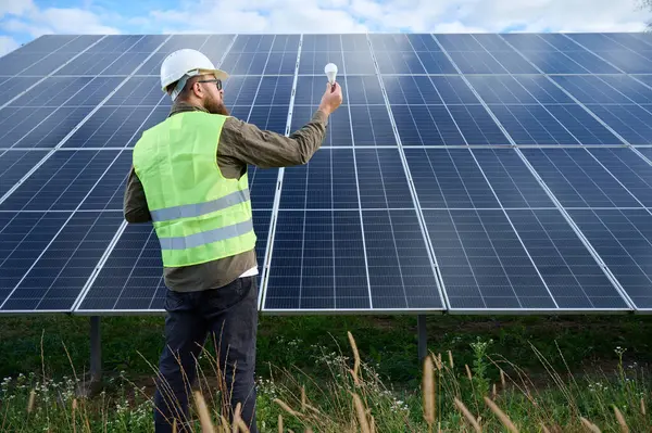 Üniformalı mühendis. Güneş istasyonu yakınlarında koruyucu kasklı. Elinde ampul var. Denetmen güneş çiftliğindeki güneş panellerini kontrol ediyor. Güneş enerjisi ve yenilenebilir enerji. Ön manzara. Yeşil enerji kavramı.