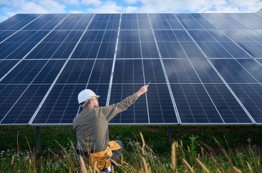 Güneş istasyonu yakınında koruyucu kasklı, üniformalı bir mühendis elinde tornavida tutuyor. Denetmen güneş çiftliğindeki güneş panellerini kontrol ediyor. Yeşil enerji kavramı.