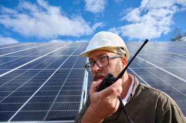 Engineer in uniform with protective helmet near solar station holding walkie talkie. Supervisor is checking on solar panels with radio station on solar farm. Solar and renewable energy. Close up clipart