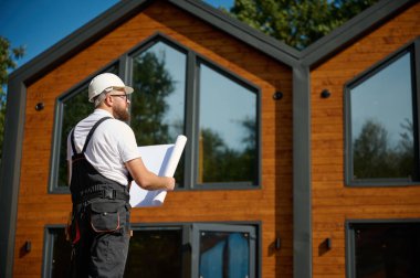 Mimar bekleme planı. Sakallı mimar elinde proje planları, yeni modern evlerin yanında duruyor ve inşaat alanında bir şeyler arıyor..