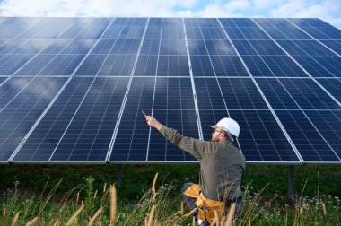Güneş istasyonu yakınında koruyucu kasklı, üniformalı bir mühendis elinde tornavida tutuyor. Denetmen güneş çiftliğindeki güneş panellerini kontrol ediyor. Yeşil enerji kavramı.