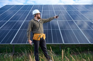 Güneş istasyonu yakınında koruyucu kasklı üniformalı bir mühendis telsiz tutuyor. Denetmen güneş panellerini güneş çiftliğindeki radyo istasyonuyla kontrol ediyor. Güneş ve yenilenebilir enerji kavramı.