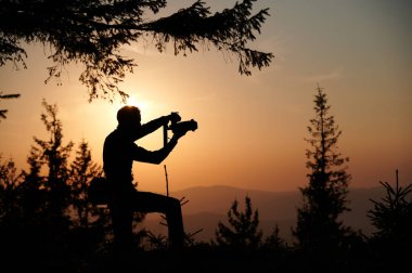 Silhouette of a photographer in the mountains taking pictures at dawn. Tourist holding camera on the mountain hill on sunrise background. Man takes photo of mountain in the rays of the setting sun. clipart