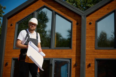 Mimar bekleme planı. Sakallı mimar elinde proje planları, yeni modern evlerin yanında duruyor ve inşaat alanında bir şeyler arıyor..