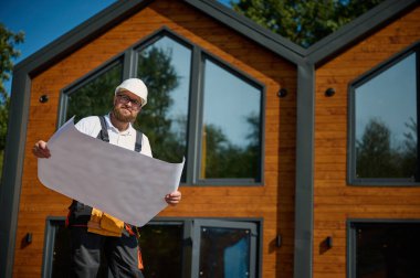 Mimar bekleme planı. Sakallı mimar elinde proje planları, yeni modern evlerin yanında duruyor ve inşaat alanında bir şeyler arıyor..