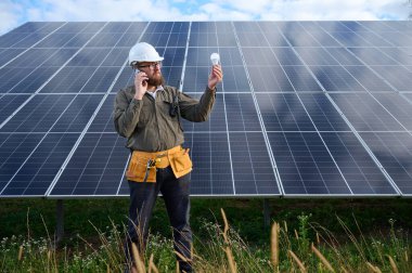 Üniformalı mühendis. Güneş istasyonu yakınlarında koruyucu kasklı. Elinde ampul var. Denetmen güneş çiftliğindeki güneş panellerini kontrol ediyor. Güneş enerjisi ve yenilenebilir enerji. Ön manzara. Yeşil enerji kavramı.