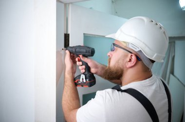 Kapı monte eden tornavidayla inşaat işçisi. Montaj için aksesuarlar, mobilya monte etmek, evi onarmak için. İş kıyafeti, kask ve koruyucu gözlük takmış bir adam. Ev yenileme konsepti