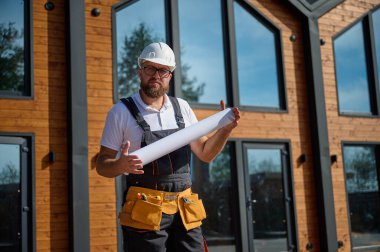 Mimar bekleme planı. Sakallı mimar elinde proje planları, yeni modern evlerin yanında duruyor ve inşaat alanında bir şeyler arıyor..