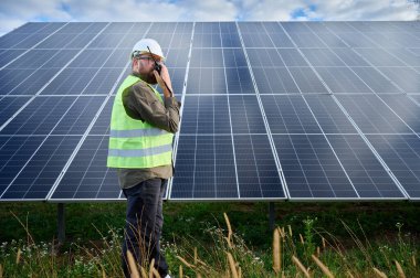 Güneş istasyonu yakınında koruyucu kasklı üniformalı bir mühendis telsiz tutuyor. Denetmen güneş panellerini güneş çiftliğindeki radyo istasyonuyla kontrol ediyor. Güneş ve yenilenebilir enerji kavramı.