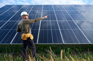 Güneş istasyonu yakınında koruyucu kasklı, üniformalı bir mühendis elinde tornavida tutuyor. Denetmen güneş çiftliğindeki güneş panellerini kontrol ediyor. Yeşil enerji kavramı.