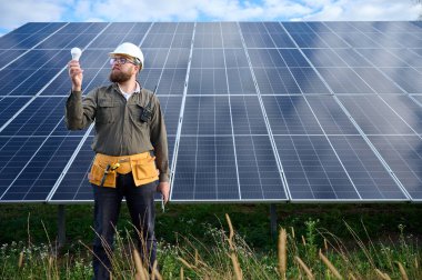 Üniformalı mühendis. Güneş istasyonu yakınlarında koruyucu kasklı. Elinde ampul var. Denetmen güneş çiftliğindeki güneş panellerini kontrol ediyor. Güneş enerjisi ve yenilenebilir enerji. Ön manzara. Yeşil enerji kavramı.