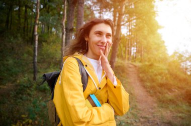 Sarı yağmurluk ve sırt çantasıyla ormanda yürüyen güzel bir kadın. Parlak giysiler içinde sonbahar yürüyüşü. Dağ yolculuğu ve açık hava eğlencesi..
