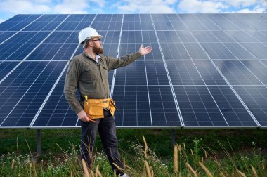 Güneş istasyonu yakınında koruyucu kasklı, üniformalı bir mühendis elinde tornavida tutuyor. Denetmen güneş çiftliğindeki güneş panellerini kontrol ediyor. Yeşil enerji kavramı.