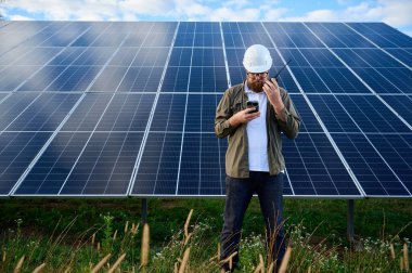Güneş istasyonu yakınında koruyucu kasklı üniformalı bir mühendis akıllı telefon ve telsiz tutuyor. Denetmen güneş panellerini güneş çiftliğindeki akıllı telefonuyla kontrol ediyor. Güneş ve yenilenebilir enerji