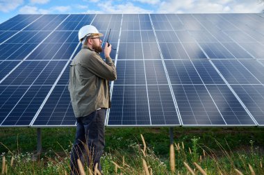 Güneş istasyonu yakınında koruyucu kasklı üniformalı bir mühendis telsiz tutuyor. Denetmen güneş panellerini güneş çiftliğindeki radyo istasyonuyla kontrol ediyor. Güneş ve yenilenebilir enerji kavramı.