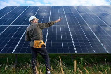 Güneş istasyonu yakınında koruyucu kasklı, üniformalı bir mühendis elinde tornavida tutuyor. Denetmen güneş çiftliğindeki güneş panellerini kontrol ediyor. Yeşil enerji kavramı.