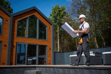 Architect hold plan. Bearded architect wearing hardhat holding project blueprints paper plans standing near comlex of new modern houses looking to something at construction site. clipart