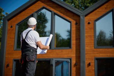 Mimar bekleme planı. Sakallı mimar elinde proje planları, yeni modern evlerin yanında duruyor ve inşaat alanında bir şeyler arıyor..