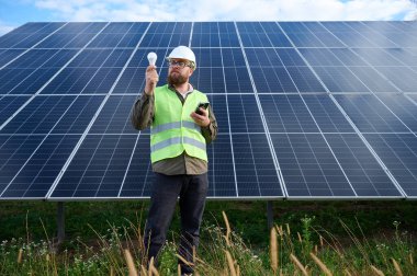 Üniformalı mühendis. Güneş istasyonu yakınlarında koruyucu kasklı. Elinde ampul var. Denetmen güneş panellerini güneş çiftliğindeki akıllı telefonuyla kontrol ediyor. Güneş ve yenilenebilir enerji kavramı.