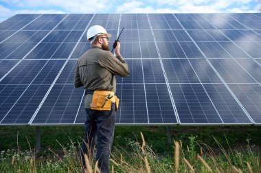 Güneş istasyonu yakınında koruyucu kasklı üniformalı bir mühendis telsiz tutuyor. Denetmen güneş panellerini güneş çiftliğindeki radyo istasyonuyla kontrol ediyor. Güneş ve yenilenebilir enerji kavramı.