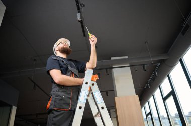 Adam yeni ofisin tavanına bir spot LED ışığı yerleştirdi. Elektrikçi merdivenden inmiyor. Sanayi tipi siyah çatı katı tasarımı. Sakallı adam yeni daireye ışık takıyor.