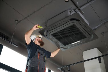 Sakallı adam merdivende duruyor ofise klima yerleştiriyor. Modern binada tavana monte edilmiş klima. Çatı katı tarzı. Endüstriyel iç tasarım. Teknisyen servisi.