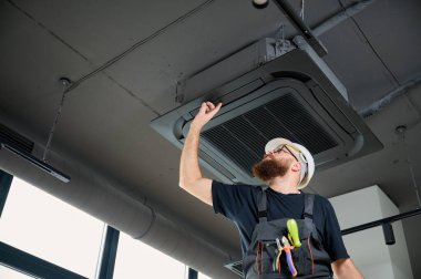 Sakallı adam merdivende duruyor ofise klima yerleştiriyor. Modern binada tavana monte edilmiş klima. Çatı katı tarzı. Endüstriyel iç tasarım. Teknisyen servisi.