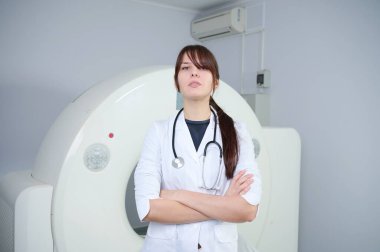 MRI tarayıcısının önünde duran kendinden emin kadın teknisyenin portresi. Beyaz laboratuvar önlüklü kadın tomografinin yanında duruyor. Tarama odasında güzel bir doktor var. Üniformalı Radyolog.