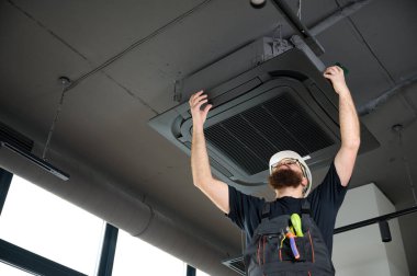 Sakallı adam merdivende duruyor ofise klima yerleştiriyor. Modern binada tavana monte edilmiş klima. Çatı katı tarzı. Endüstriyel iç tasarım. Teknisyen servisi.