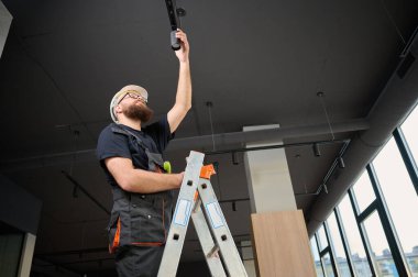 Adam yeni ofisin tavanına bir spot LED ışığı yerleştirdi. Elektrikçi merdivenden inmiyor. Sanayi tipi siyah çatı katı tasarımı. Sakallı adam yeni daireye ışık takıyor.