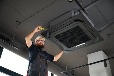 Sakallı adam merdivende duruyor ofise klima yerleştiriyor. Modern binada tavana monte edilmiş klima. Çatı katı tarzı. Endüstriyel iç tasarım. Teknisyen servisi.