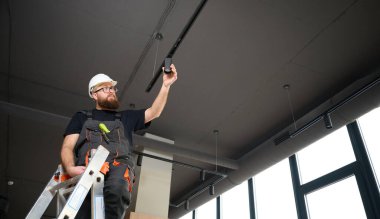 Adam yeni ofisin tavanına bir spot LED ışığı yerleştirdi. Elektrikçi merdivenden inmiyor. Sanayi tipi siyah çatı katı tasarımı. Sakallı adam yeni daireye ışık takıyor.