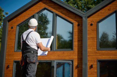 Mimar bekleme planı. Sakallı mimar elinde proje planları, yeni modern evlerin yanında duruyor ve inşaat alanında bir şeyler arıyor..