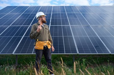 Güneş istasyonu yakınında koruyucu kasklı, üniformalı bir mühendis elinde tornavida tutuyor. Denetmen güneş çiftliğindeki güneş panellerini kontrol ediyor. Yeşil enerji kavramı.