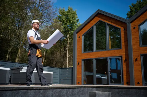 Mimar bekleme planı. Sakallı mimar elinde proje planları, yeni modern evlerin yanında duruyor ve inşaat alanında bir şeyler arıyor..