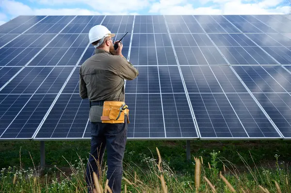 Güneş istasyonu yakınında koruyucu kasklı üniformalı bir mühendis telsiz tutuyor. Denetmen güneş panellerini güneş çiftliğindeki radyo istasyonuyla kontrol ediyor. Güneş ve yenilenebilir enerji kavramı.