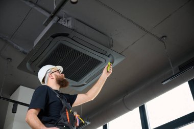 Sakallı adam merdivende duruyor ofise klima yerleştiriyor. Modern binada tavana monte edilmiş klima. Çatı katı tarzı. Endüstriyel iç tasarım. Teknisyen servisi.