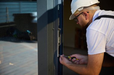 İnşaat işçisi yeni bir inşaat alanına kapı kuruyor. İş elbiseli adam ön kapıyı kuruyor. Üniformalı müteahhit tornavidayla yeni güzel evin ön cam kapısını ayarlıyor..