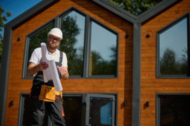 Mimar bekleme planı. Sakallı mimar elinde proje planları, yeni modern evlerin yanında duruyor ve inşaat alanında bir şeyler arıyor..