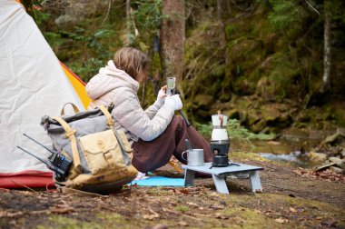 Güzel bir blogcu kız ormanda çadırın yanında oturur ve akıllı telefon kamerasıyla kendini filme çeker. Gezgin blogcu kadın kamp çadırının yakınında sosyal video hikayesi yazıyor. açık hava vlog kaydedicisi