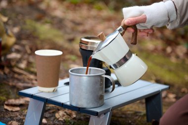 Açık hava kahve seti piknik masasında ve ormanda kamp. Kamp masasında Moka pot kahve, kamp. Kahve ocağı doğası. Bardağı kahveyle dolduran kadın.