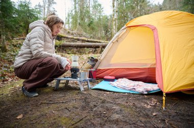 Yürüyüşçü kadın kamp çadırının yanında kahve hazırlıyor. İlkbaharın başlarında ormanda bir bayan gezgin sıcak kahveyi bir turist bardağında tutuyor..