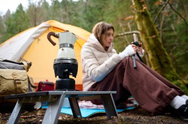Ormanda çadırın yanında oturan bir kadın kahve sehpasıyla elinde akıllı telefonuyla. Macera, seyahat, turizm ve kamp konsepti.