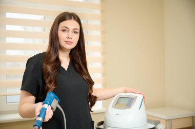 Klinikte tıbbi kıyafet giymiş genç bir kız rehabilitasyon uzmanı elinde şok dalgası terapisi için bir cihaz tutuyor. Yaralardan sonra iyileşme kavramı. Extracorporeal Shockwave Terapisi