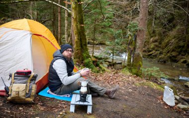 Kamp çadırının yanında oturan ve elinde sıcak kahveyle bir kupa tutan bir adam. Macera, seyahat, turizm ve kamp konsepti. Kahve içen bir adam doğada kamp yapmaktan hoşlanıyor..