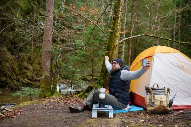 Kamp çadırının yakınında takılan bir adam. Macera, seyahat, turizm ve kamp konsepti. Adam doğada kamp yaparken kahve içiyor. Kampın yakınında uzanan adam.