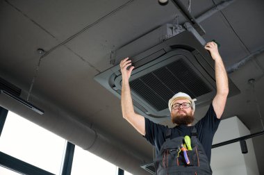 Sakallı adam merdivende duruyor ofise klima yerleştiriyor. Modern binada tavana monte edilmiş klima. Çatı katı tarzı. Endüstriyel iç tasarım. Teknisyen servisi.