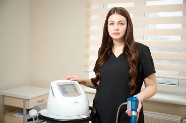 Klinikte tıbbi kıyafet giymiş genç bir kız rehabilitasyon uzmanı elinde şok dalgası terapisi için bir cihaz tutuyor. Yaralardan sonra iyileşme kavramı. Extracorporeal Shockwave Terapisi